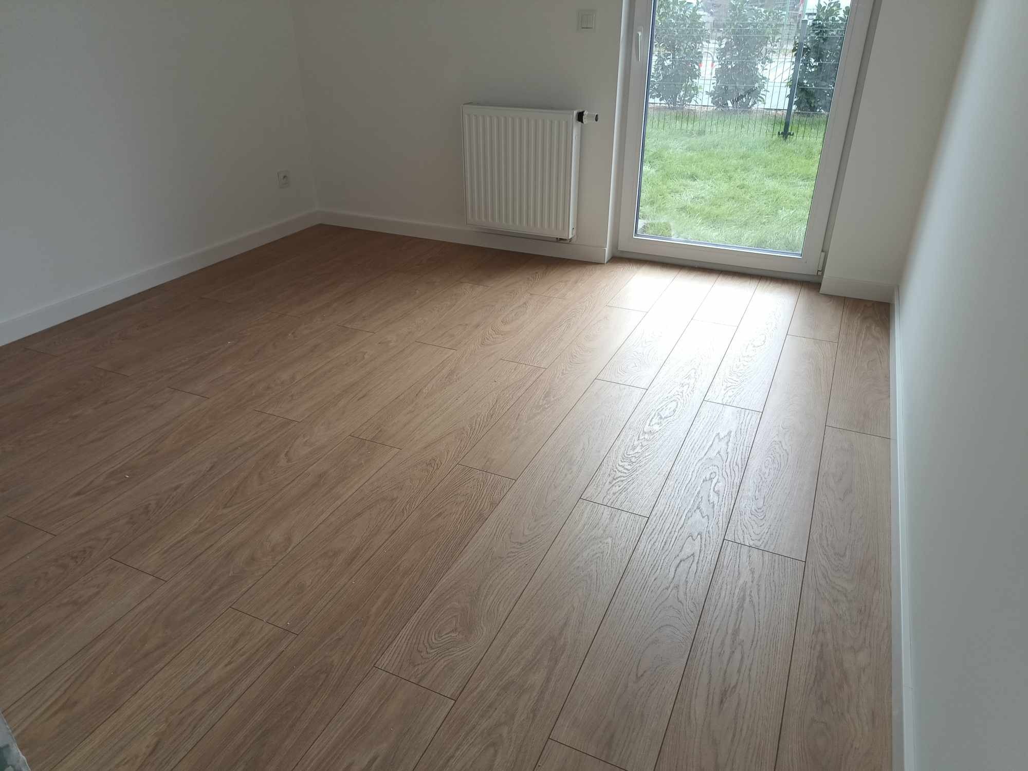 Jasne panele podłogowe ułożone w nowym pokoju z grzejnikiem i widokiem na ogród przez okno balkonowe.