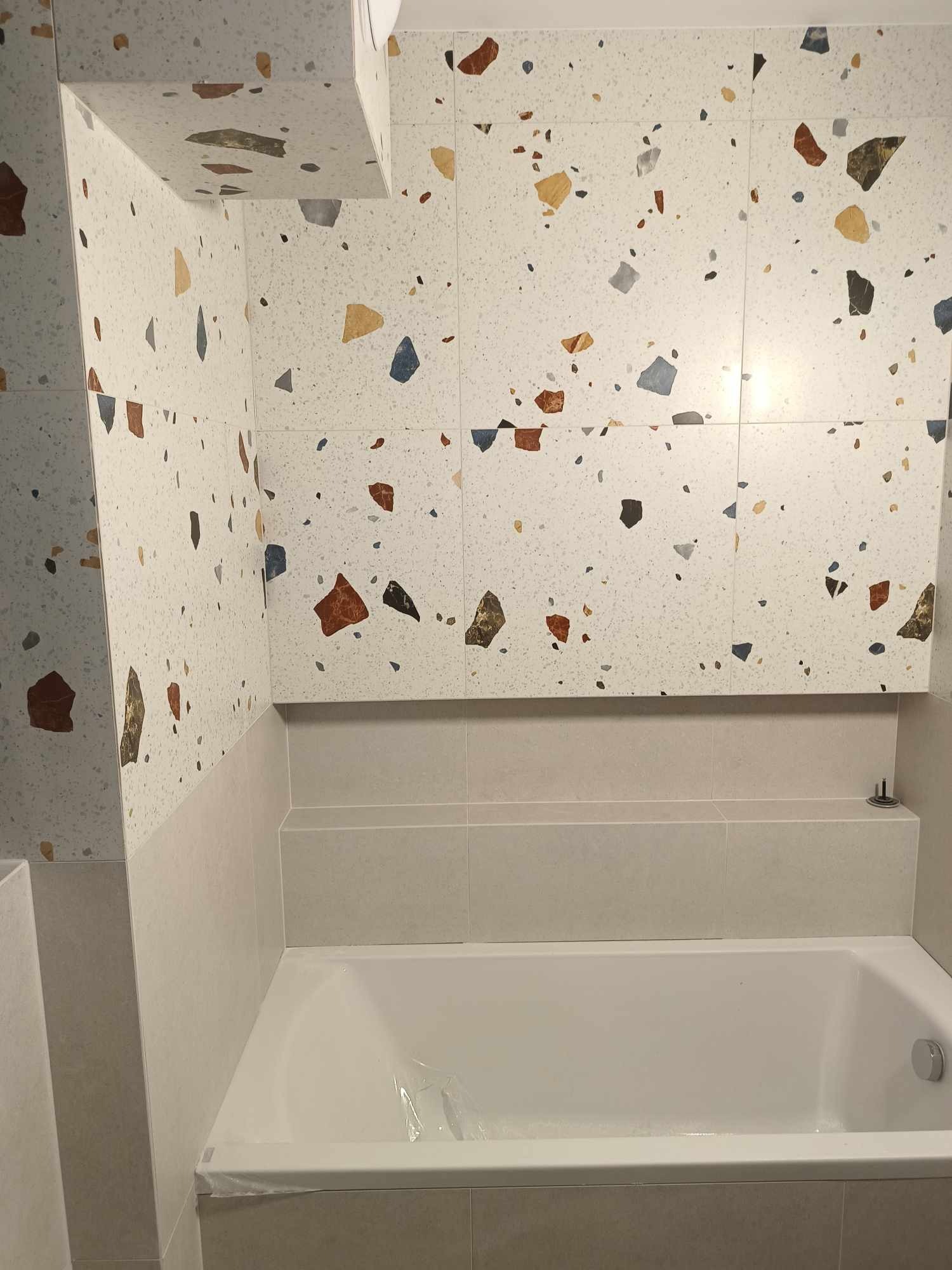 Wykończona łazienka z wanną, ściany i zabudowa wanny wyłożone płytkami imitującymi lastryko w odcieniach bieli, beżu, brązu i niebieskiego, widoczne zabezpieczenie folią na wannie.