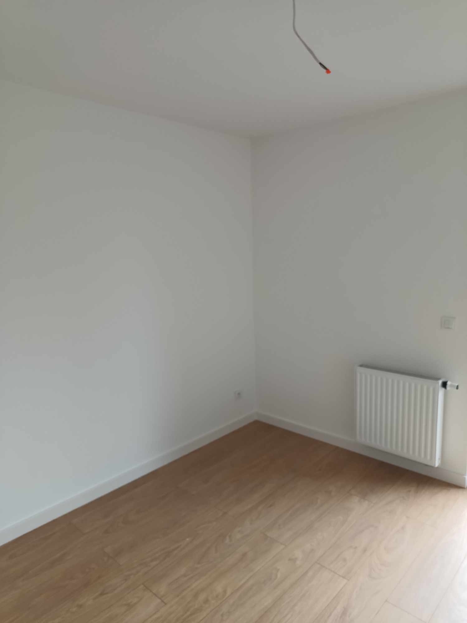 Pusty, odświeżony pokój z panelami podłogowymi w odcieniach jasnego drewna, białymi ścianami, grzejnikiem i wystającą ze ściany instalacją elektryczną.