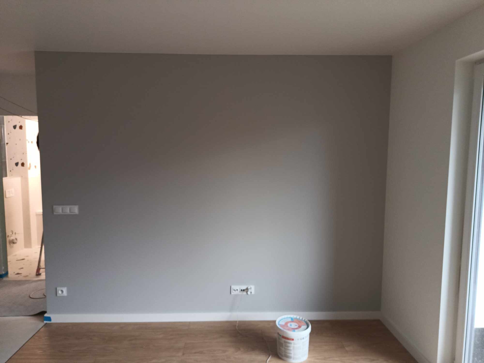 Świeżo pomalowana szara ściana w pokoju z drewnianą podłogą; widoczna puszka farby, gniazdka elektryczne i listwy przypodłogowe.
