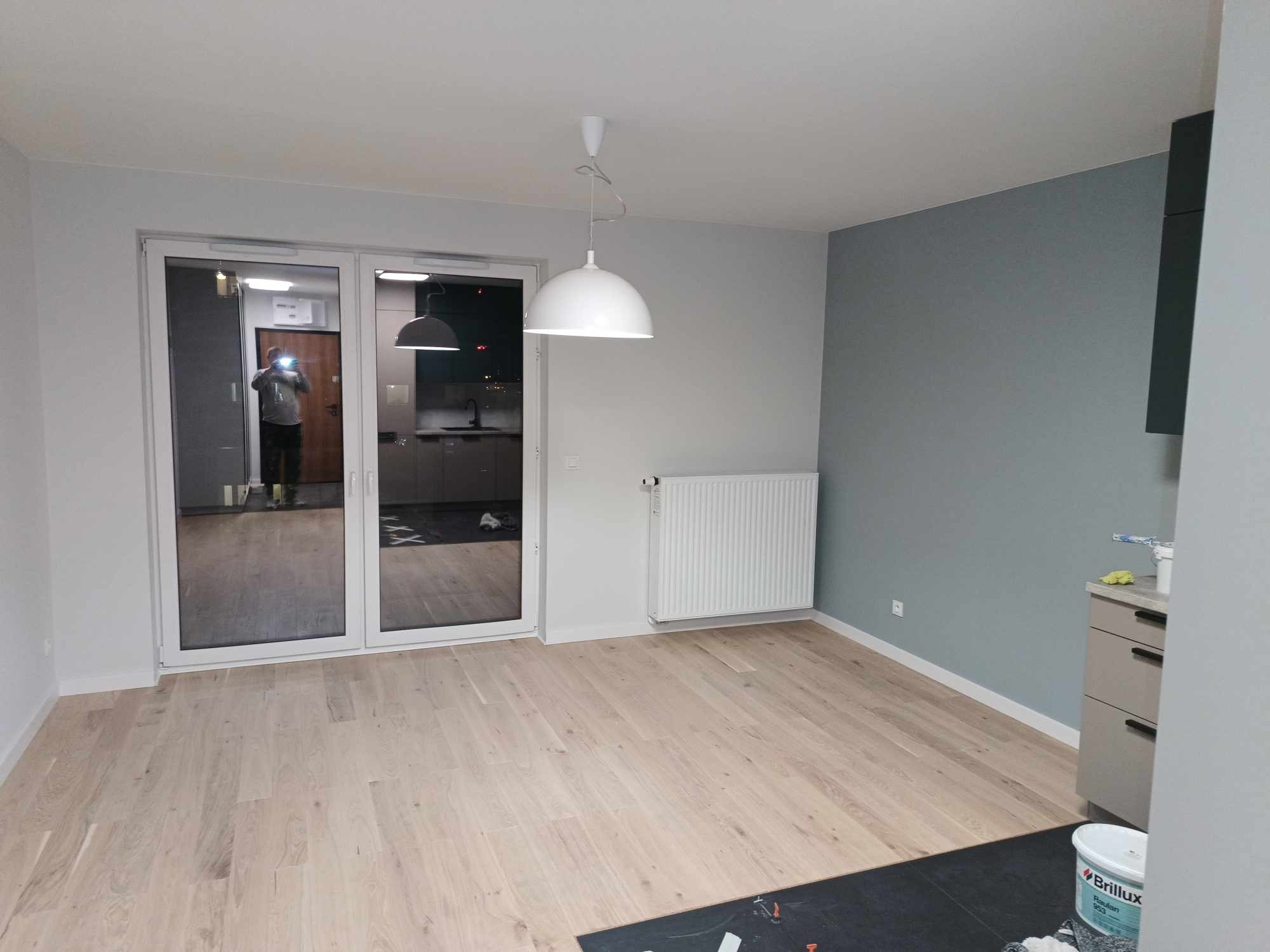 Jasne drewniane panele podłogowe w nowo wyremontowanym pokoju z szarą ścianą, białym grzejnikiem i dużymi przesuwnymi drzwiami balkonowymi, przez które widać wyposażoną kuchnię. Na podłodze leżą...