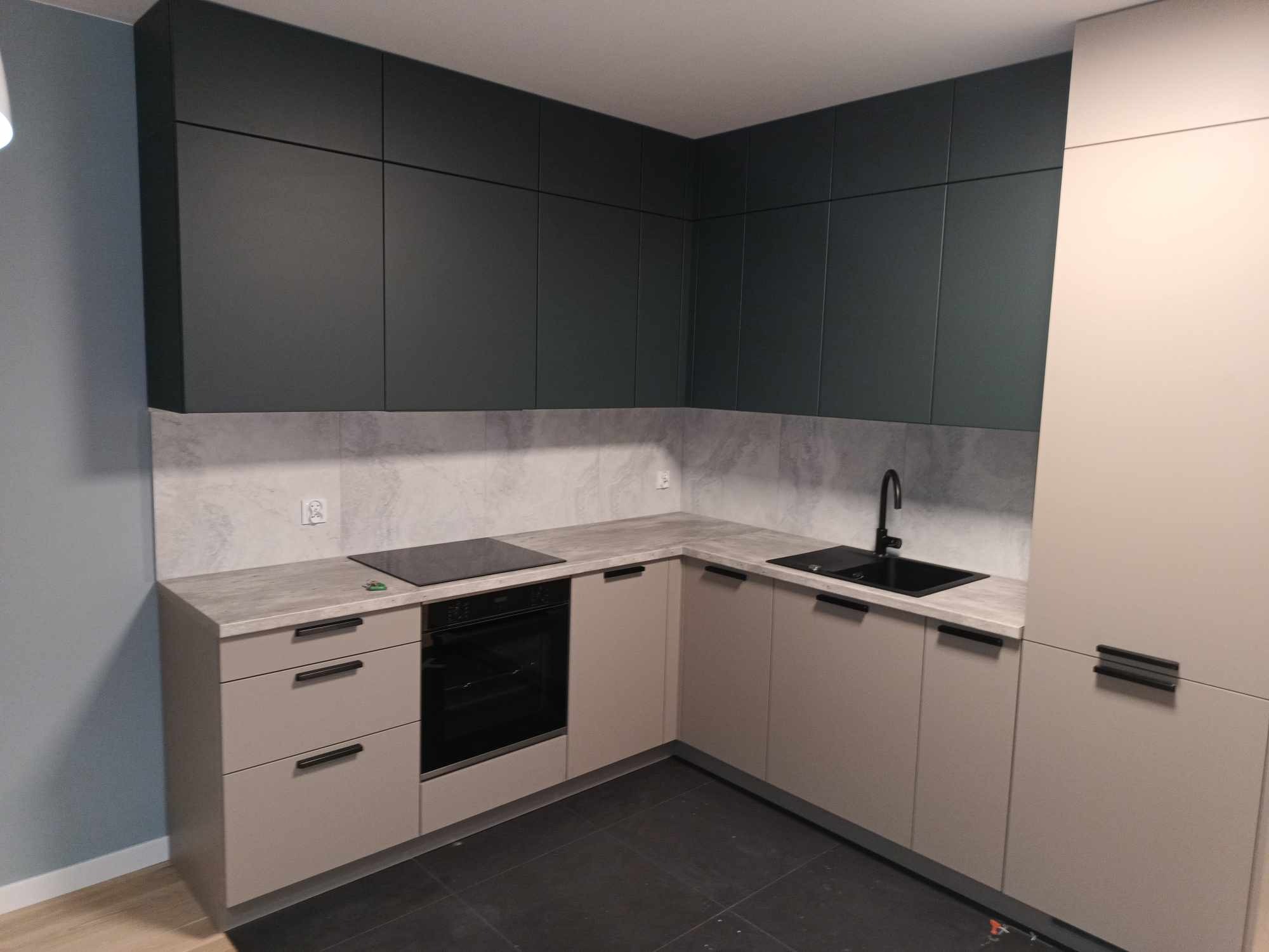 Nowoczesna kuchnia w odcieniach beżu i grafitu z czarną armaturą i płytą indukcyjną, minimalistyczne fronty szafek bez uchwytów, szara mozaika na ścianie nad blatem, czarna podłoga z płytek.