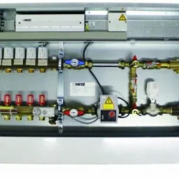 Rozdzielacz ogrzewania podłogowego w metalowej obudowie, widoczne liczne zawory, rury miedziane i regulatory, całość zamontowana w schludnej szafce.