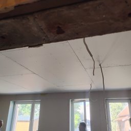 Ryn-Bud - Sufit z płyt kartonowo-gipsowych, widoczne łączenia płyt i wystające przewody elektryczne, fragment drewnianej konstrukcji stropu w górnej części kadru, okna w tle.