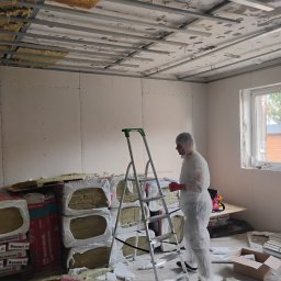 Ryn-Bud - Pracownik w kombinezonie ochronnym stoi przy drabinie w pomieszczeniu z zamontowanym stelażem sufitowym i częściowo ocieplonym stropem, obok leżą paczki wełny mineralnej Rockwool i worki z zaprawą...