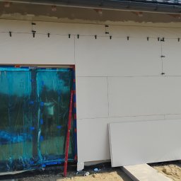 Ryn-Bud - Ocieplanie elewacji budynku styropianem, okno zabezpieczone folią, poziomica oparta o ścianę, widoczne mocowania paneli.