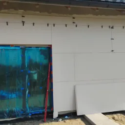 Ocieplanie elewacji budynku styropianem, okno zabezpieczone folią, poziomica oparta o ścianę, widoczne mocowania paneli.