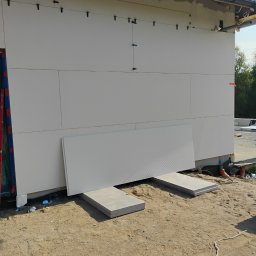 Ryn-Bud - Fragment ocieplonej ściany budynku z zamocowanymi płytami izolacyjnymi, z widocznymi elementami mocującymi oraz leżącą płytą na betonowych podkładach.