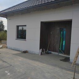 Ryn-Bud - Nowoczesny dom jednorodzinny z jasną elewacją w trakcie wykończenia, widoczne panele elewacyjne, okno zabezpieczone folią, oraz fragment wnętrza z drewnianymi panelami ściennymi i narzędziami.