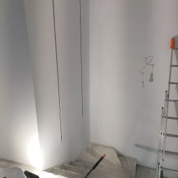 Ryn-Bud - Wnętrze w trakcie malowania: białe ściany, schody betonowe, wiadro z farbą, wałek malarski, drabina aluminiowa i lampa halogenowa oświetlająca ścianę.