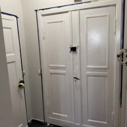 Ryn-Bud - Białe, dwuskrzydłowe drzwi w trakcie malowania, zabezpieczone niebieską taśmą malarską przy ścianach i framugach, widoczne ślady świeżej farby i odsłonięte gniazdko elektryczne.