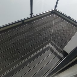 Ryn-Bud - Nowoczesny balkon z szarymi deskami kompozytowymi i szklaną balustradą z czarnymi ramami, widok z góry z fragmentem buta i szmatki na podłodze.