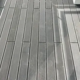 Szare deski kompozytowe tworzące podłogę balkonu, ułożone równolegle, z widocznymi drobnymi zabrudzeniami i refleksami światła.