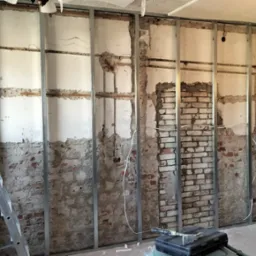 Remont pokoju: montaż metalowego stelażu pod płytę karton gips na tle odsłoniętej ceglanej ściany, widoczna drabina i narzędzia.