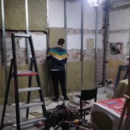 Ryn-Bud - Pracownik montuje izolację z wełny mineralnej w metalowym stelażu na ścianie z odsłoniętą cegłą, widoczne narzędzia na stole, drabina i resztki starego tynku na suficie.
