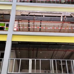 Ryn-Bud - Wnętrze budynku w trakcie prac wykończeniowych, widoczne surowe ściany, instalacje, zabezpieczenia taśmami i przeszklony sufit.