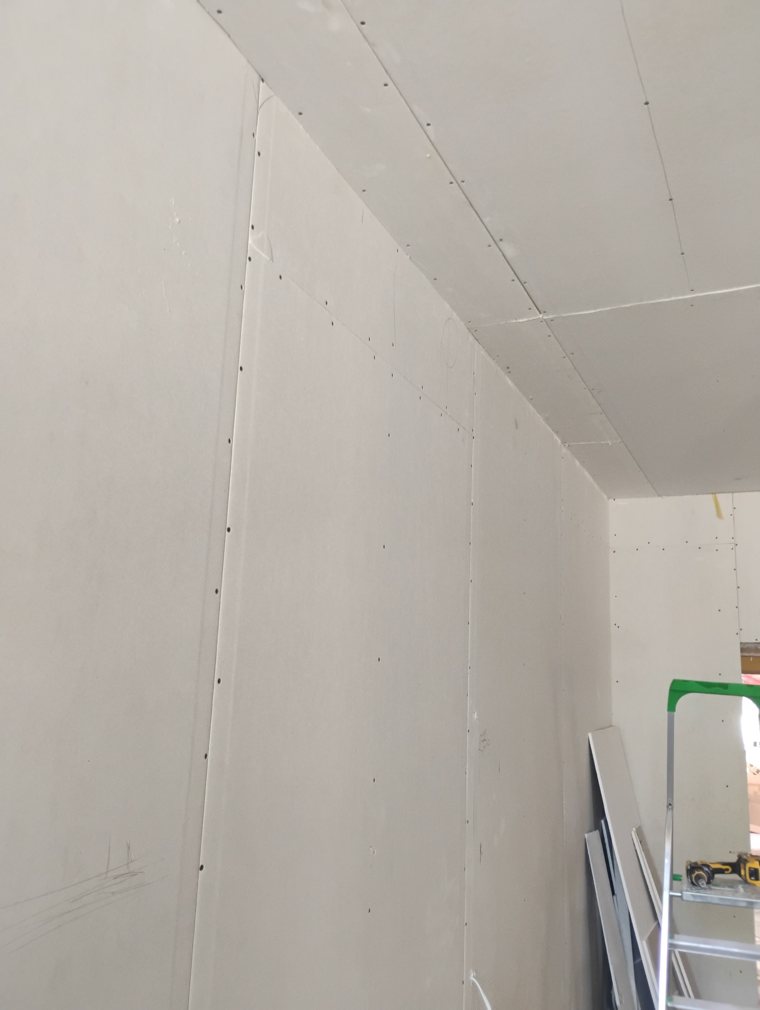 Ściana i sufit pokryte płytami kartonowo-gipsowymi, widoczne łączenia i śruby, w tle drabina i narzędzia.