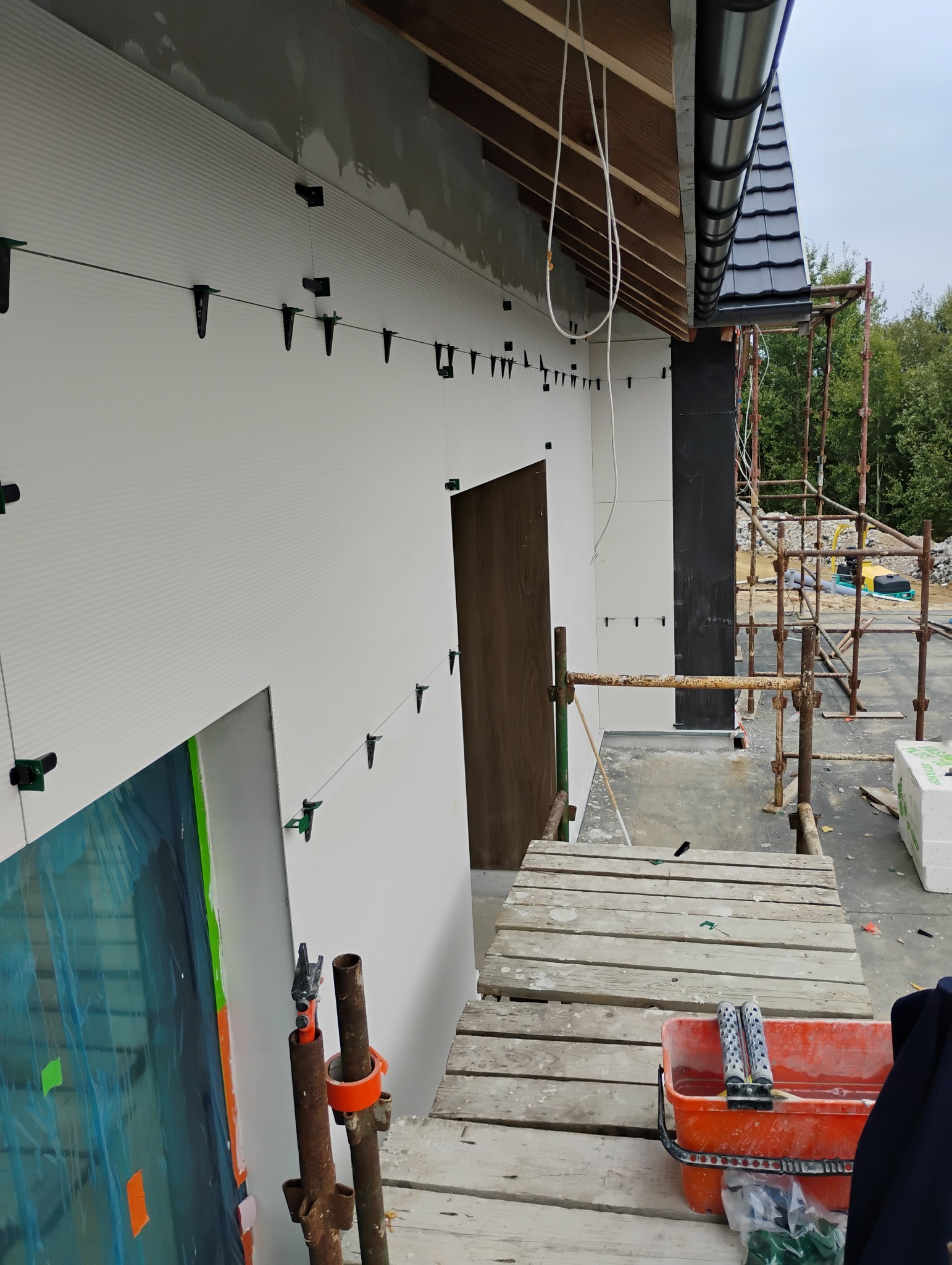 Ocieplanie elewacji budynku z widocznymi elementami mocującymi styropian i rusztowaniem w tle.