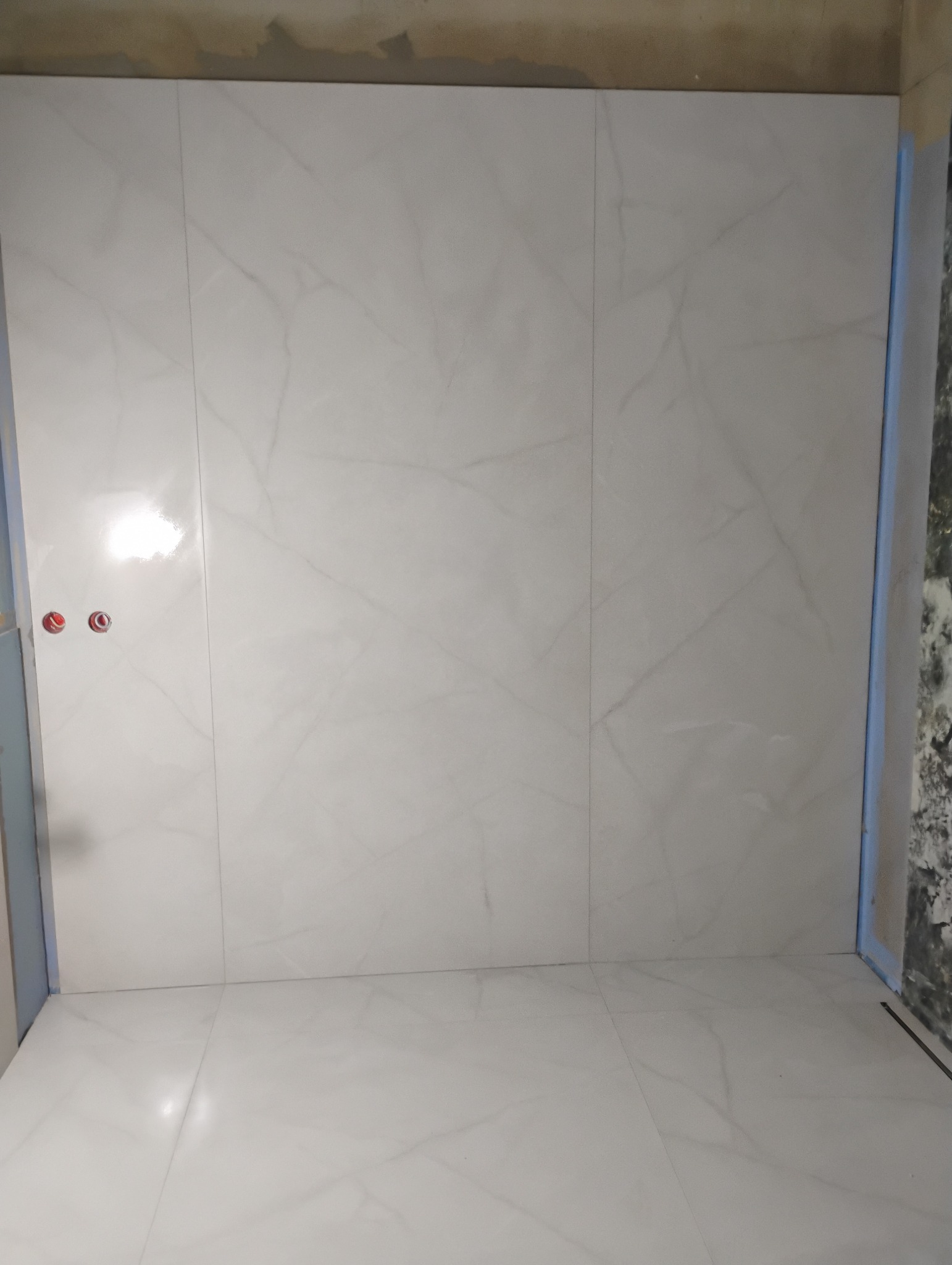 Wyłożona białymi płytkami imitującymi marmur ściana i podłoga w trakcie remontu łazienki. Widoczne wystające rury instalacji wodnej oraz zabezpieczone krawędzie.