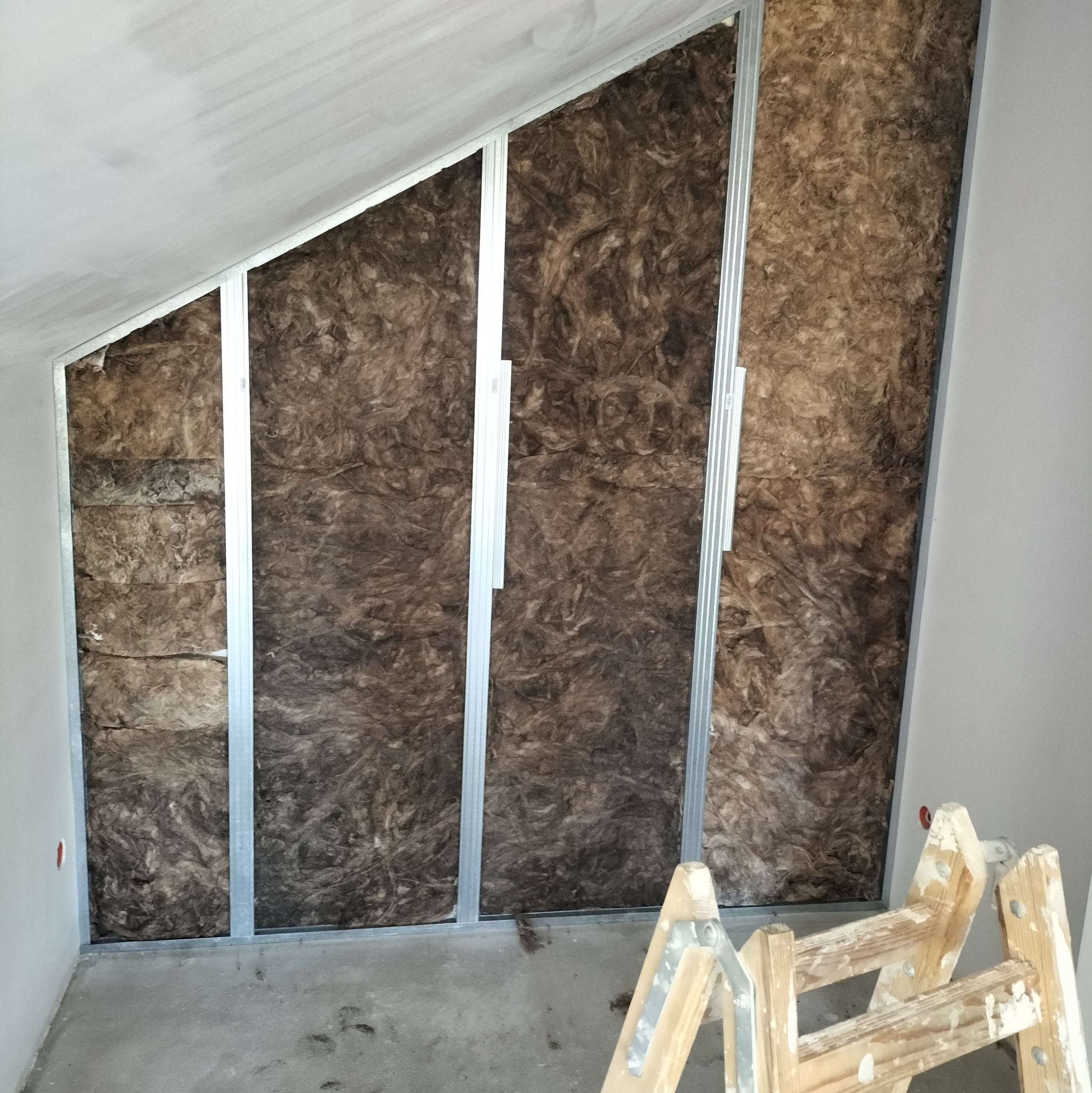 Skośna ściana poddasza z zamontowanym rusztem metalowym i wypełniona brązową wełną mineralną, widoczna drabina malarska.