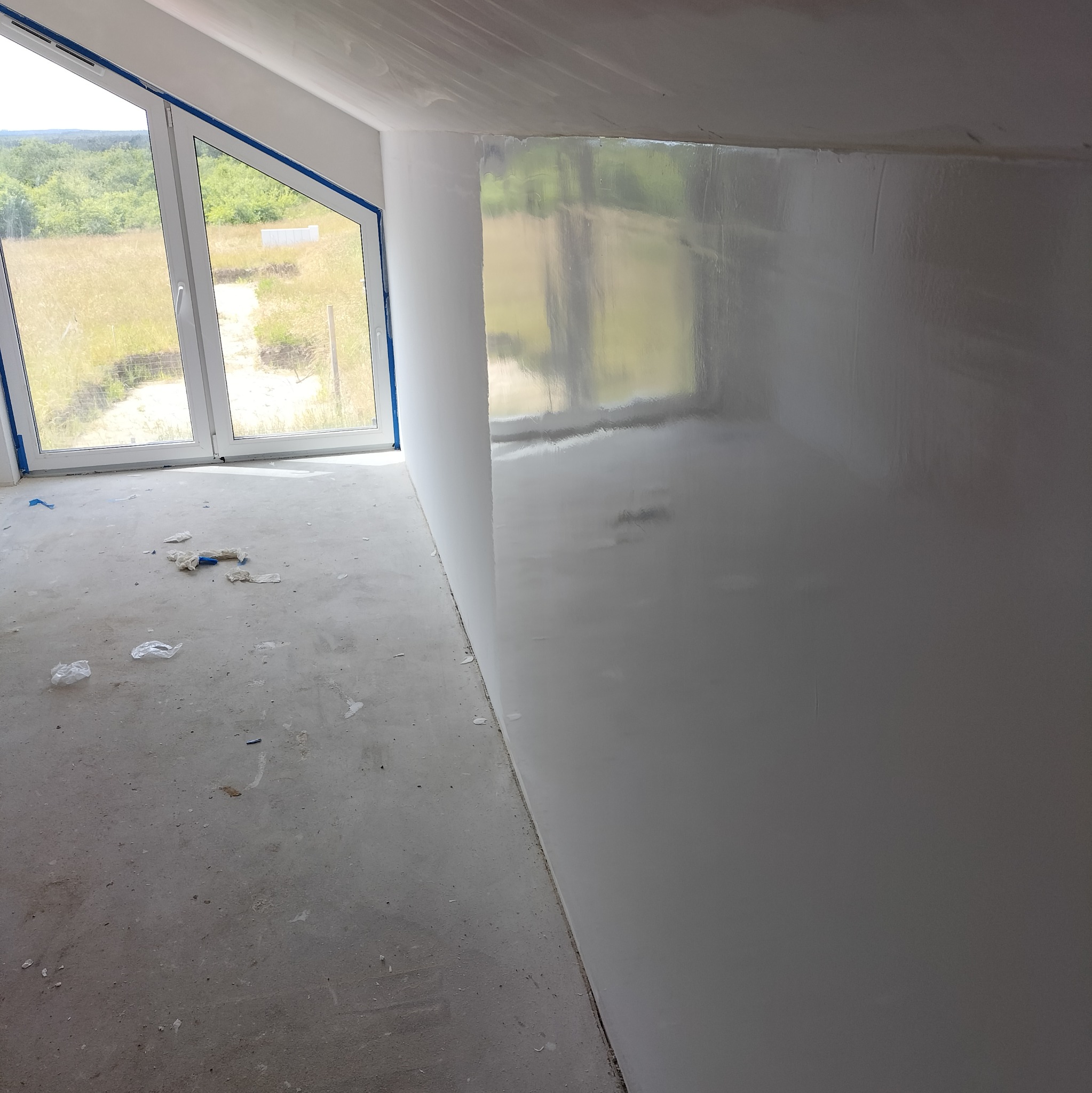 Świeżo wygładzona ściana w pokoju na poddaszu z oknem, zabezpieczona folią sufit, na podłodze resztki materiałów budowlanych.