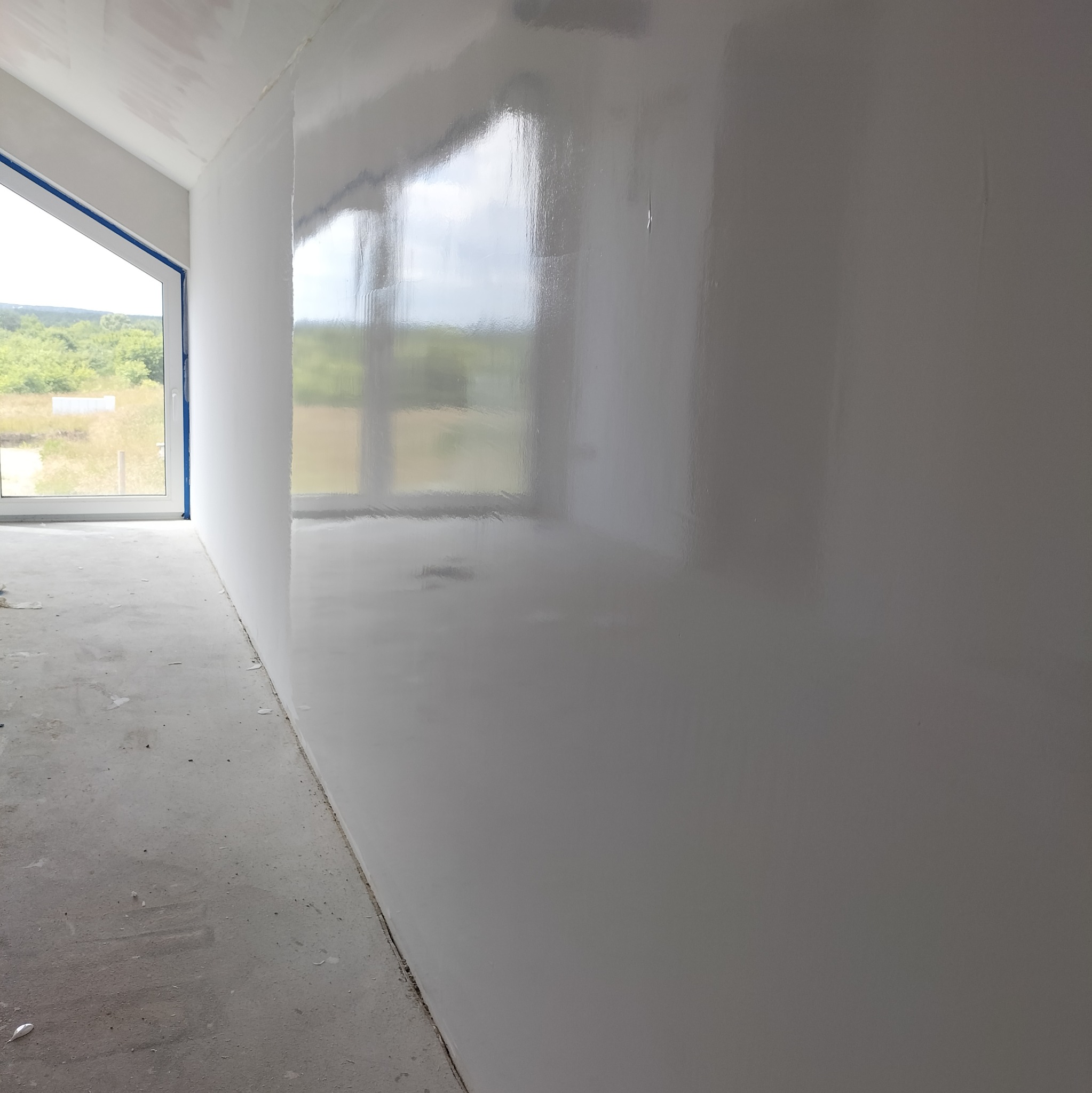 Świeżo położona, błyszcząca gładź na ścianie w pokoju z oknem, widoczne refleksy światła, zabezpieczone taśmą malarską ramy okienne, surowy beton na podłodze.