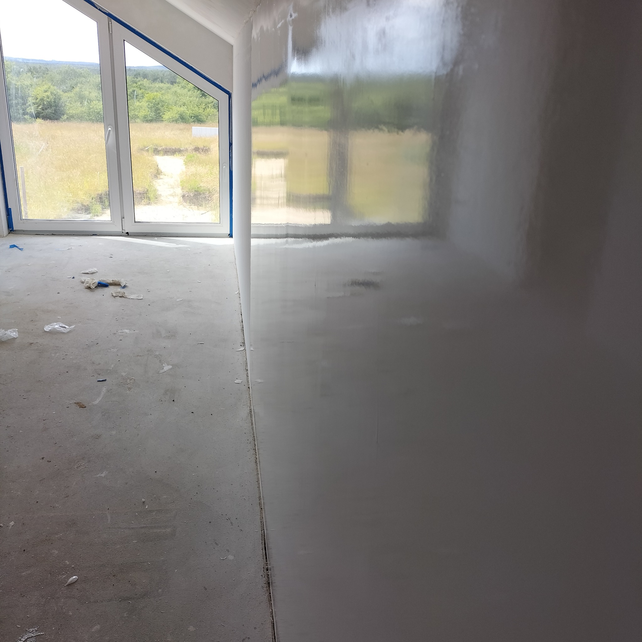 Świeżo nałożona, błyszcząca gładź na ścianie w surowym wnętrzu z oknem z widokiem na zewnątrz, widoczne ślady po pracach wykończeniowych na podłodze.