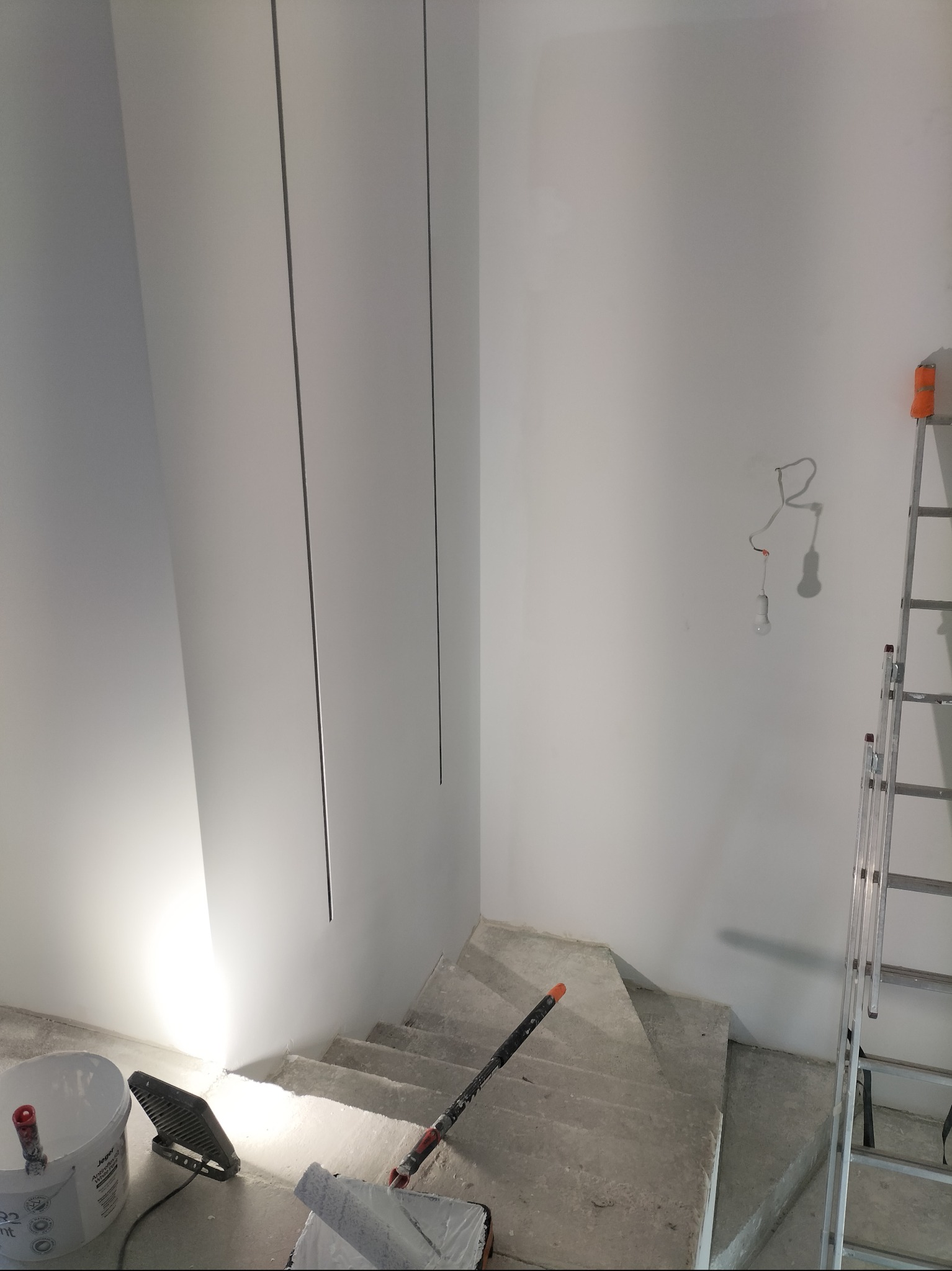 Wnętrze w trakcie malowania: białe ściany, schody betonowe, wiadro z farbą, wałek malarski, drabina aluminiowa i lampa halogenowa oświetlająca ścianę.