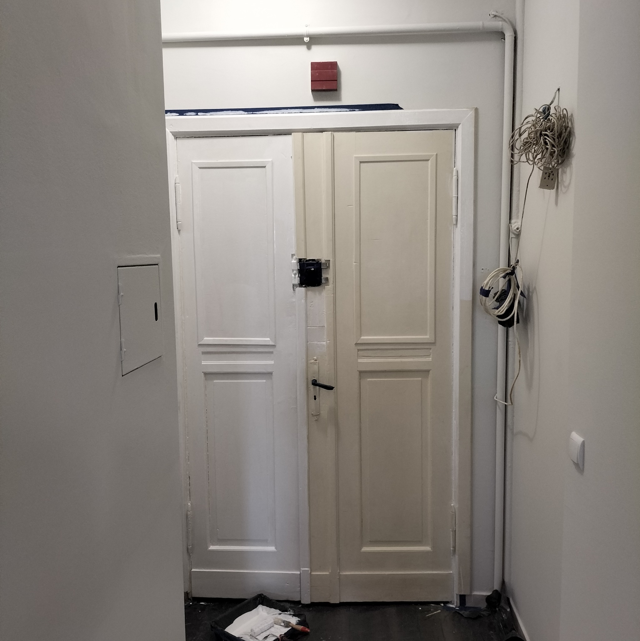 Dwudrzwiowe drzwi pomalowane w połowie na biało, z widocznym wiadrem farby i wałkiem u dołu, nieuporządkowane kable zwisające ze ściany obok rury.