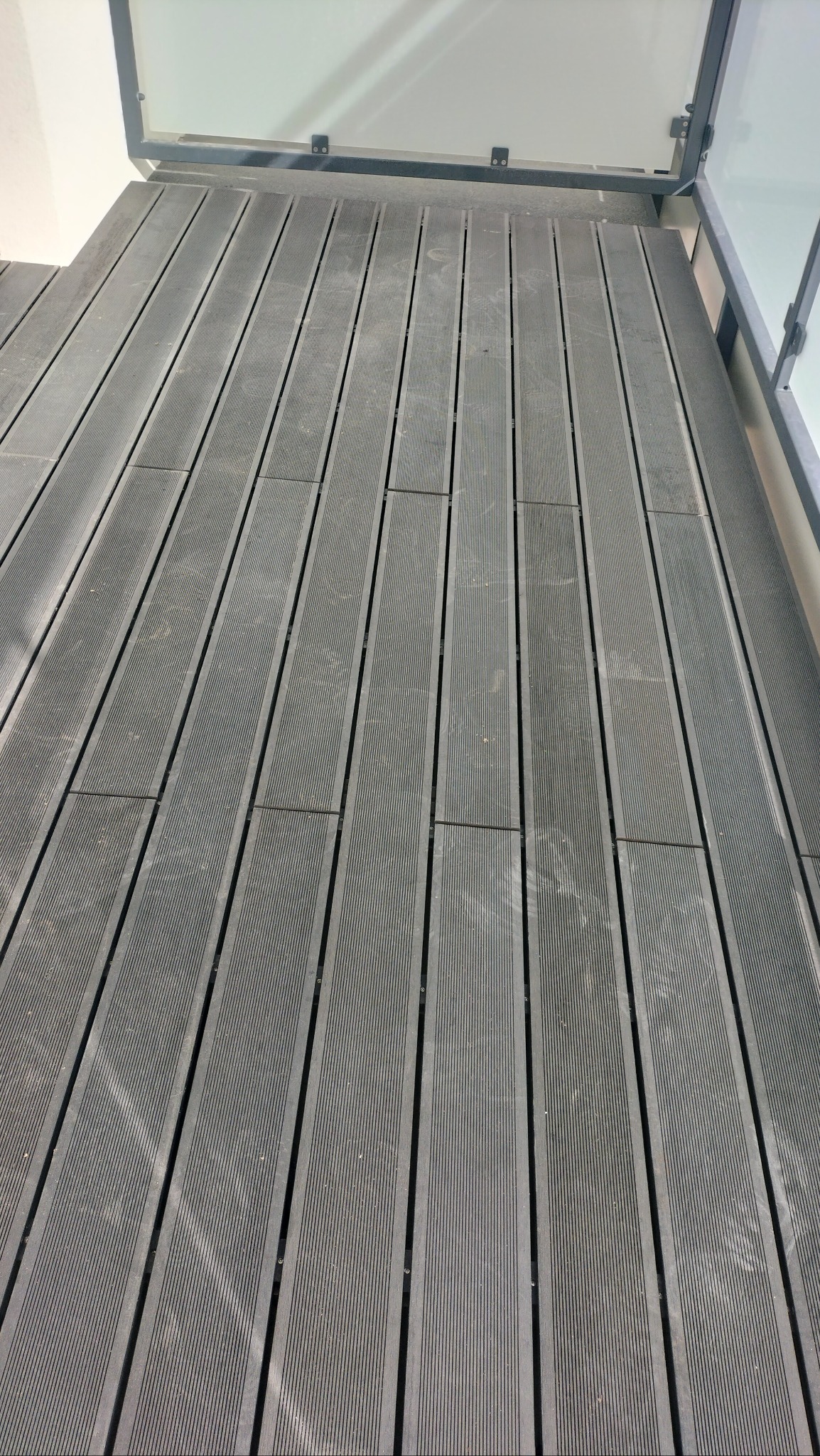 Szare deski kompozytowe tworzące taras, widoczne ślady użytkowania i fragment szklanej balustrady.