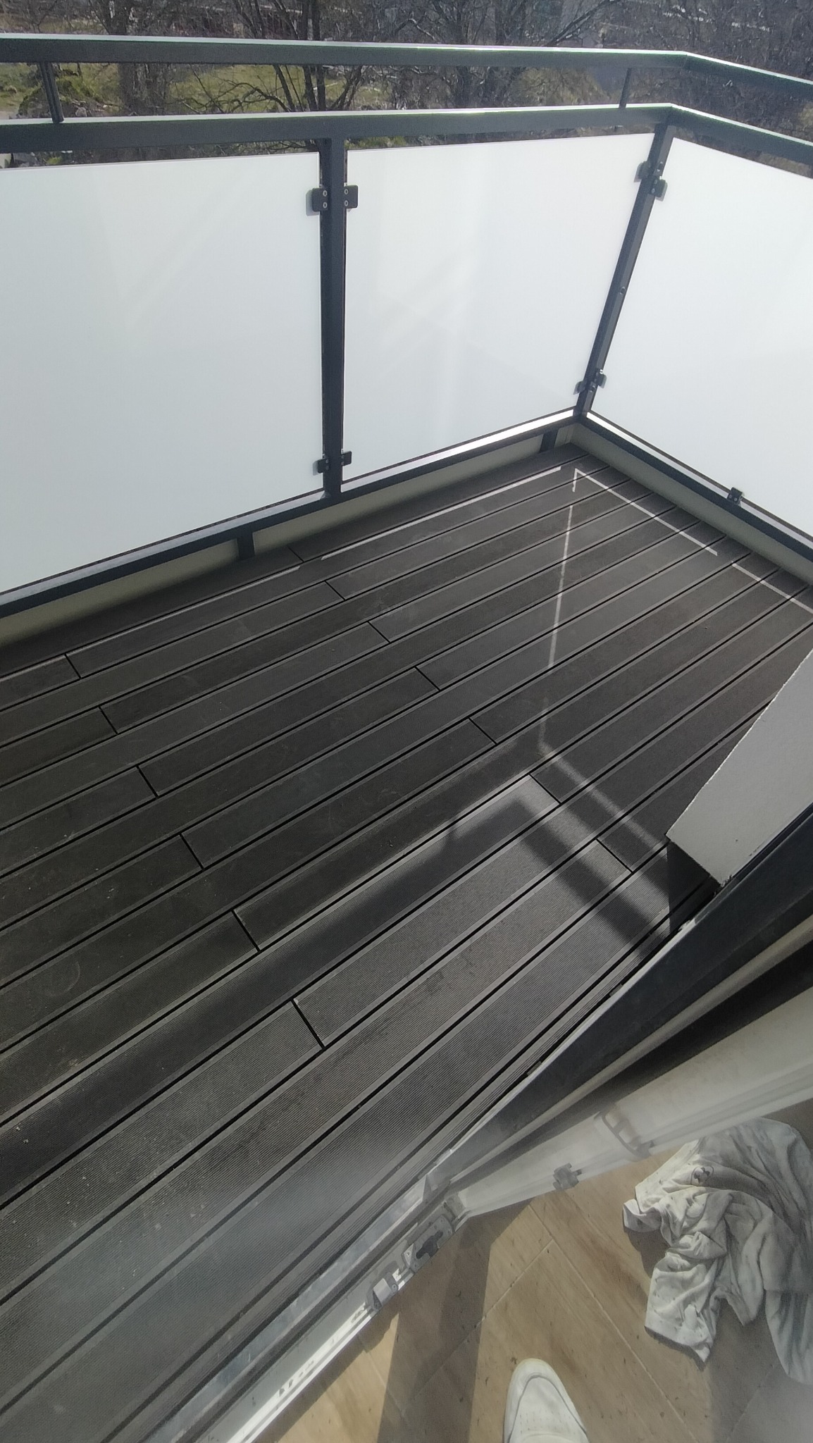 Nowoczesny balkon z szarymi deskami kompozytowymi i szklaną balustradą z czarnymi ramami, widok z góry z fragmentem buta i szmatki na podłodze.
