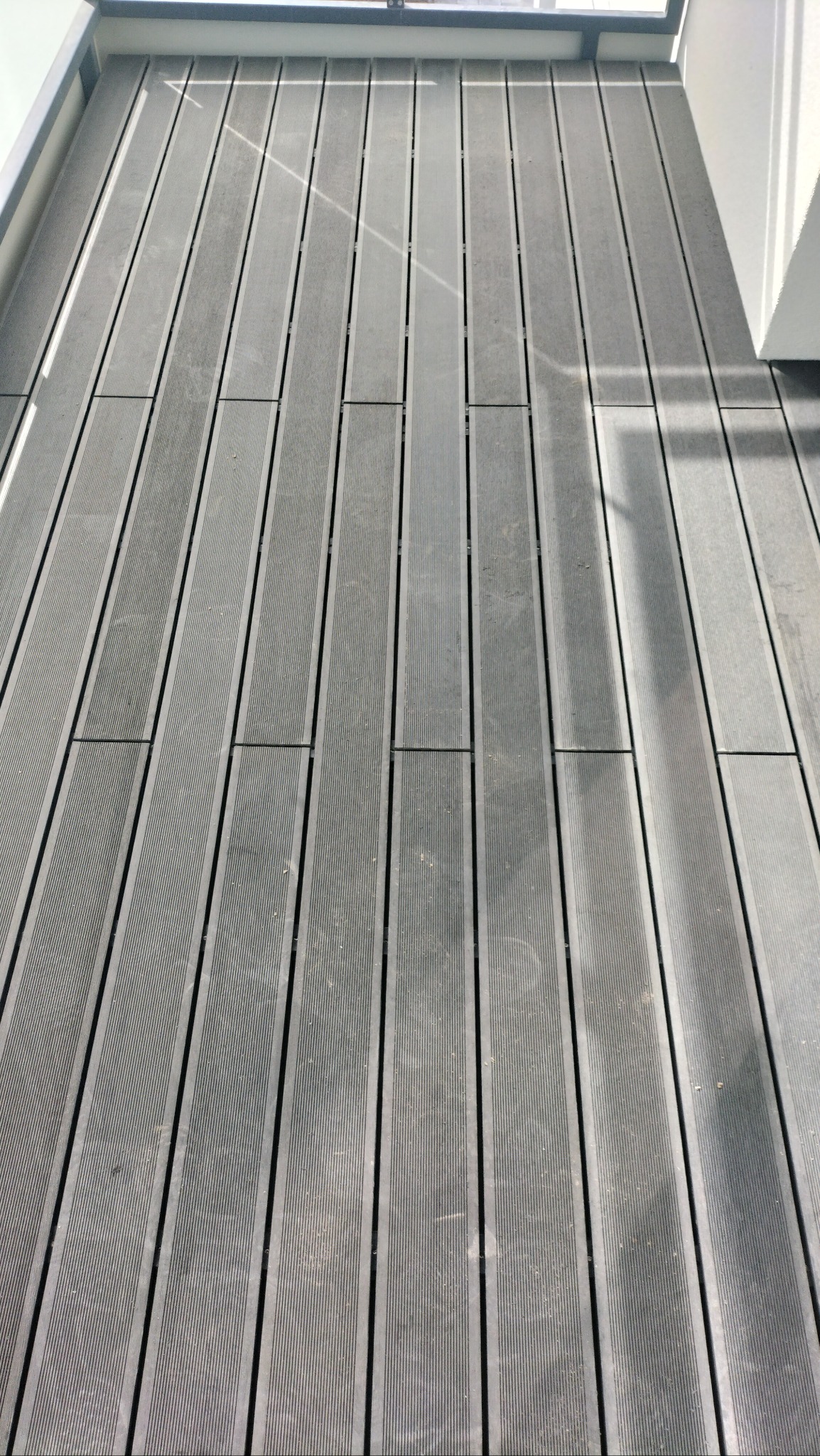 Szare deski kompozytowe tworzące podłogę balkonu, ułożone równolegle, z widocznymi drobnymi zabrudzeniami i refleksami światła.
