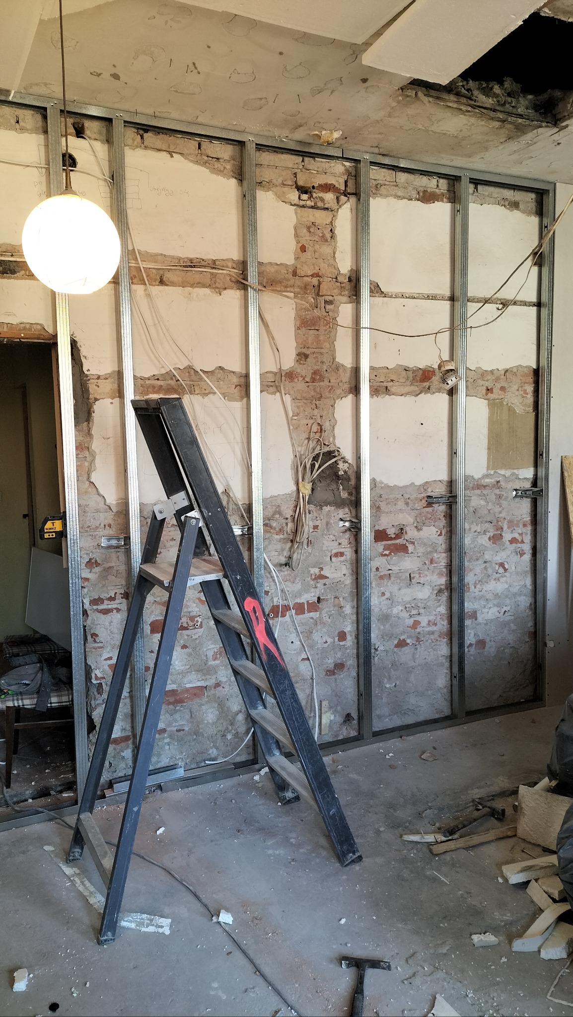 Metalowa konstrukcja szkieletowa na tle starej ceglanej ściany z resztkami tynku, drabina aluminiowa oparta o ścianę, widoczne instalacje elektryczne, lampa wisząca na kablu, fragment sufitu...