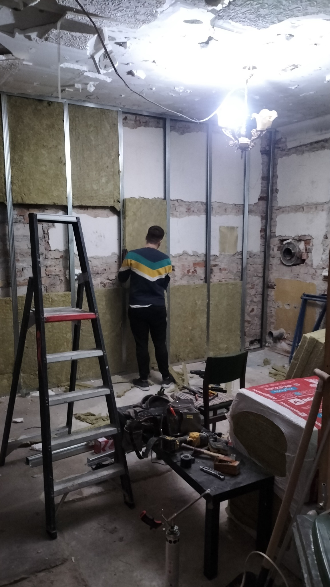 Pracownik montuje izolację z wełny mineralnej w metalowym stelażu na ścianie z odsłoniętą cegłą, widoczne narzędzia na stole, drabina i resztki starego tynku na suficie.