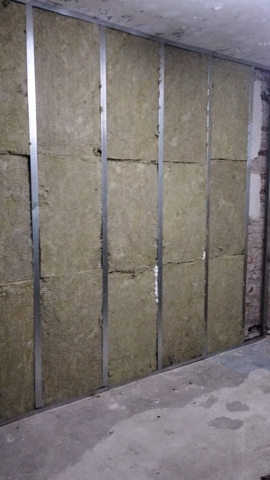 Montaż izolacji z wełny mineralnej w metalowym stelażu na ścianie, widoczne fragmenty starego muru i surowy beton na podłodze.