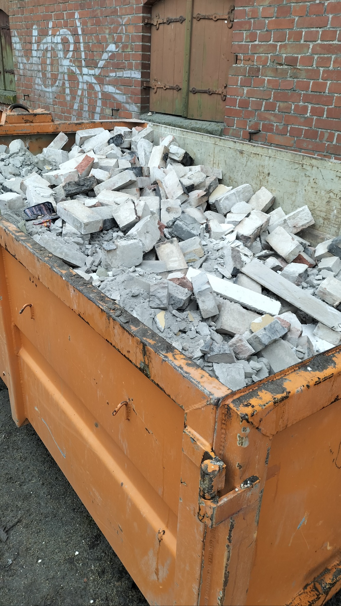 Pomarańczowy kontener wypełniony gruzem budowlanym na tle ceglanego budynku z zamurowanymi oknami.
