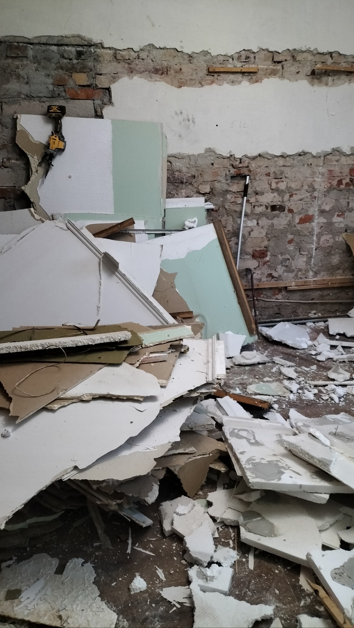 Demontaż ściany, widoczne fragmenty płyt kartonowo-gipsowych, cegieł i instalacji elektrycznej, w tle fragment ściany z odspojoną warstwą tynku, narzędzie wiertarka na ścianie.