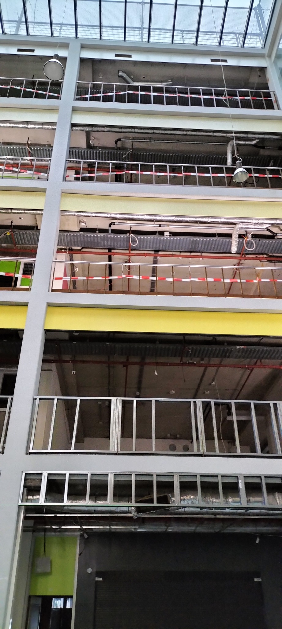 Wnętrze budynku w trakcie prac wykończeniowych, widoczne surowe ściany, instalacje, zabezpieczenia taśmami i przeszklony sufit.