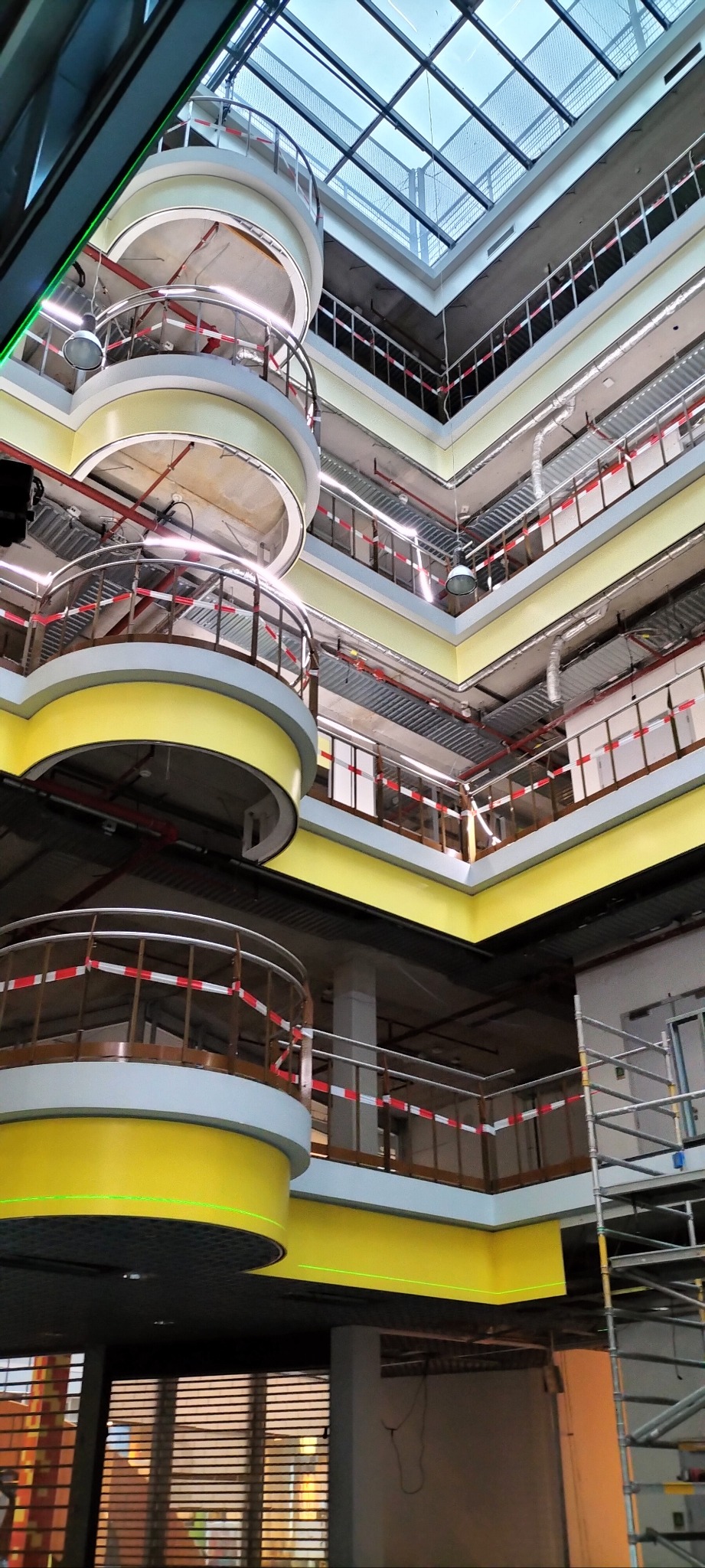 Wnętrze budynku w trakcie wykończenia, widoczne żółte elementy architektoniczne, balustrady z taśmą ostrzegawczą, sufit z przeszkleniami, rusztowania.