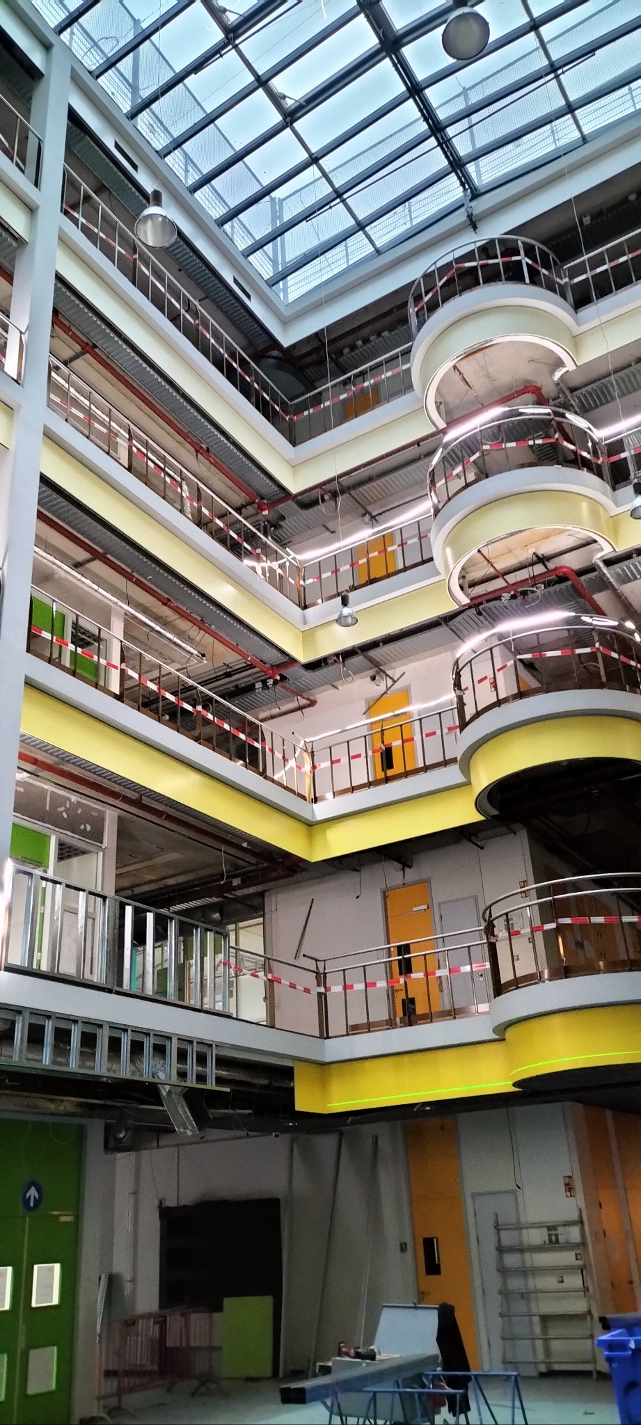 Wnętrze budynku w trakcie prac wykończeniowych z widocznymi wieloma poziomami, balkonami z żółtymi balustradami i szklanym dachem, zabezpieczone taśmami ostrzegawczymi.