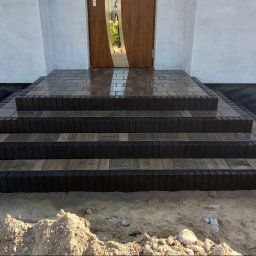 MK-BRUK Dawid Nikrant - Trzy stopnie schodów wejściowych wyłożone kostką w odcieniach szarości i brązu, z ciemnymi obrzeżami, prowadzące do drewnianych drzwi z lustrzanym panelem, widok z dołu na plac budowy.