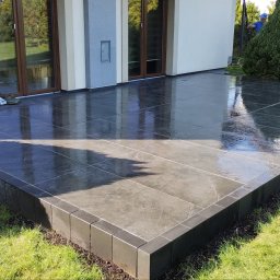 MK-BRUK Dawid Nikrant - Świeżo umyta, ciemna, kwadratowa kostka na tarasie domu, otoczona zielonym trawnikiem i ozdobnymi krzewami; widoczny pracownik w tle.