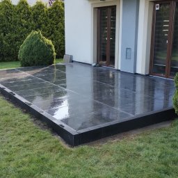 MK-BRUK Dawid Nikrant - Nowoczesny taras z ciemnych, błyszczących płyt, otoczony zielonym trawnikiem i tujami, przylegający do jasnego domu z balkonem.