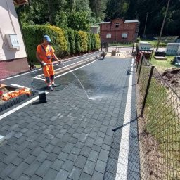 MK-BRUK Dawid Nikrant - Pracownik w pomarańczowym kombinezonie spryskuje wodą ułożoną, szarą kostkę brukową z białymi obrzeżami. W tle widoczny budynek z czerwonej cegły i zielone tuje.