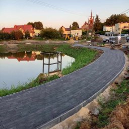 MK-BRUK Dawid Nikrant - Nowa, wijąca się ścieżka z ciemnoszarej kostki brukowej obok stawu, w tle zabudowa wiejska i kościół.