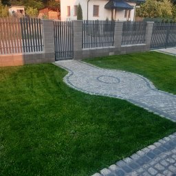 MK-BRUK Dawid Nikrant - Ułożona ścieżka z kostki granitowej z dekoracyjnym okręgiem na trawniku, otoczona nowoczesnym ogrodzeniem z metalowymi przęsłami i słupkami z betonowych bloczków.