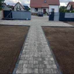 MK-BRUK Dawid Nikrant - Ułożona kostka brukowa w kształcie krzyża prowadzi do domu z szarym ogrodzeniem i bramą. Świeżo posiana trawa na rabatach. Widok z perspektywy pieszego.