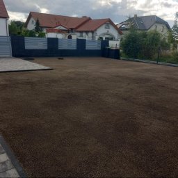 MK-BRUK Dawid Nikrant - Przygotowanie gruntu pod trawnik: wyrównany teren z widoczną bramą wjazdową i fragmentem chodnika z kostki brukowej na tle zabudowy jednorodzinnej.