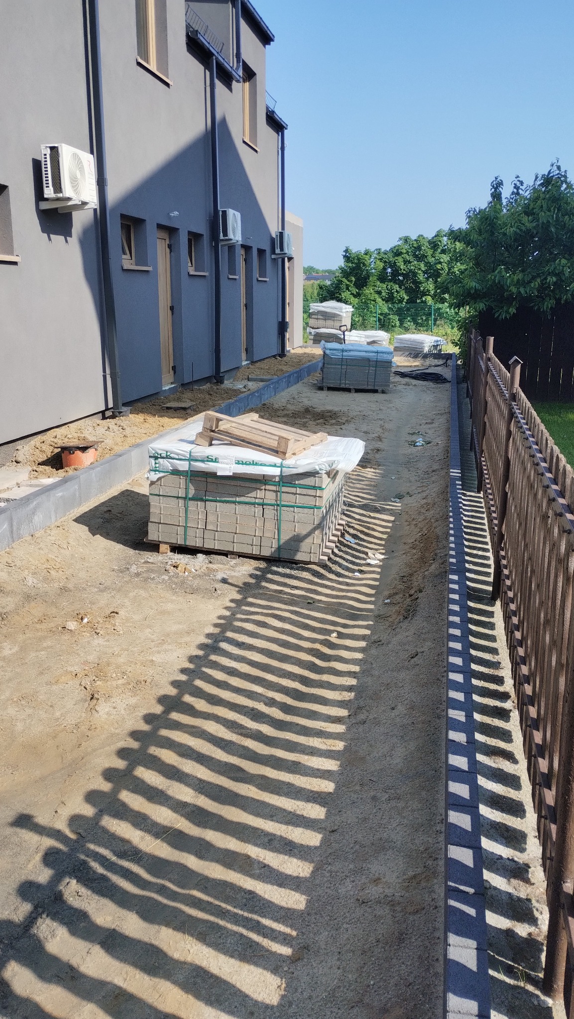 Stosy kostki brukowej na piasku obok budynku z szarą elewacją i drewnianym ogrodzeniem. Cienie rzucane przez ogrodzenie tworzą wzór na piasku.