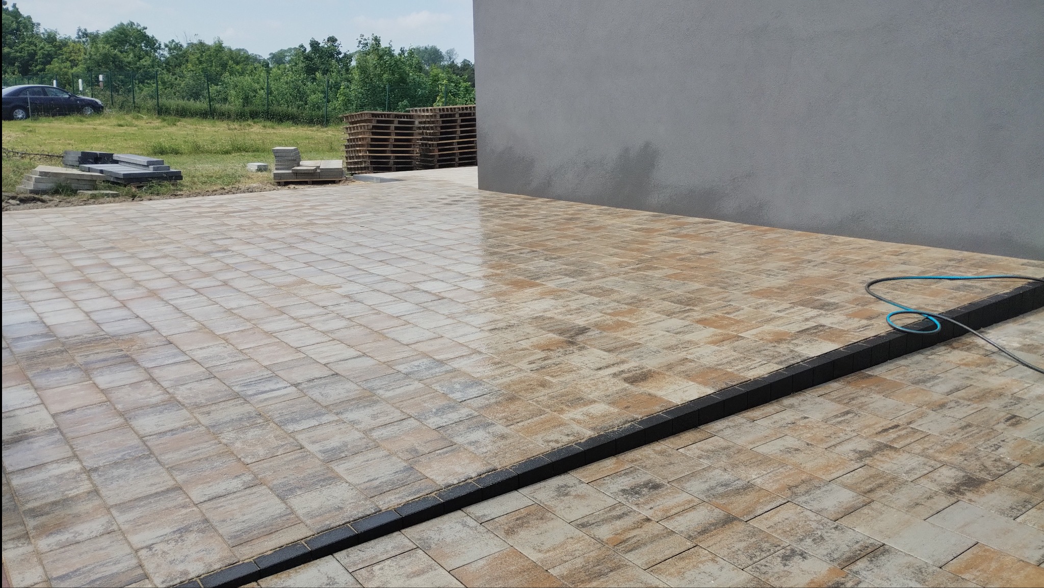 Ułożona kostka brukowa w odcieniach beżu i szarości, oddzielona czarnym obrzeżem. Część powierzchni jest mokra, prawdopodobnie po myciu. W tle budynek i zieleń.